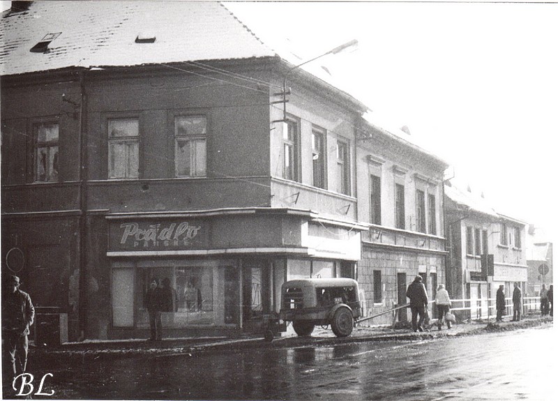 Okénko do Gottwaldovi ulice v roce 1984, tam co byl postaven Prior, dnes OD Andy. Technická záležitost - ten vozíček, kompresor ke sbíječce. Tehdy častá rekvizita stavebních prací. Dnes už vidět není. Zde stojí u obchodu s prádlem. Vývěsní štít si nechal udělat ve 30. letech módní salon Schulz a vydržel až do konce existence domu (zdroj: Českolipské obrázky)